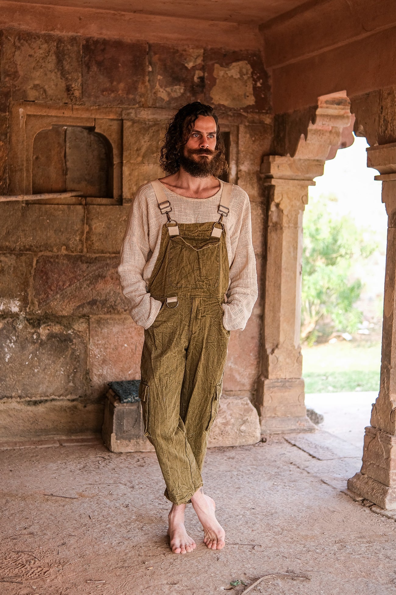 Person wearing green overalls standing in a stone-walled room with an open door.