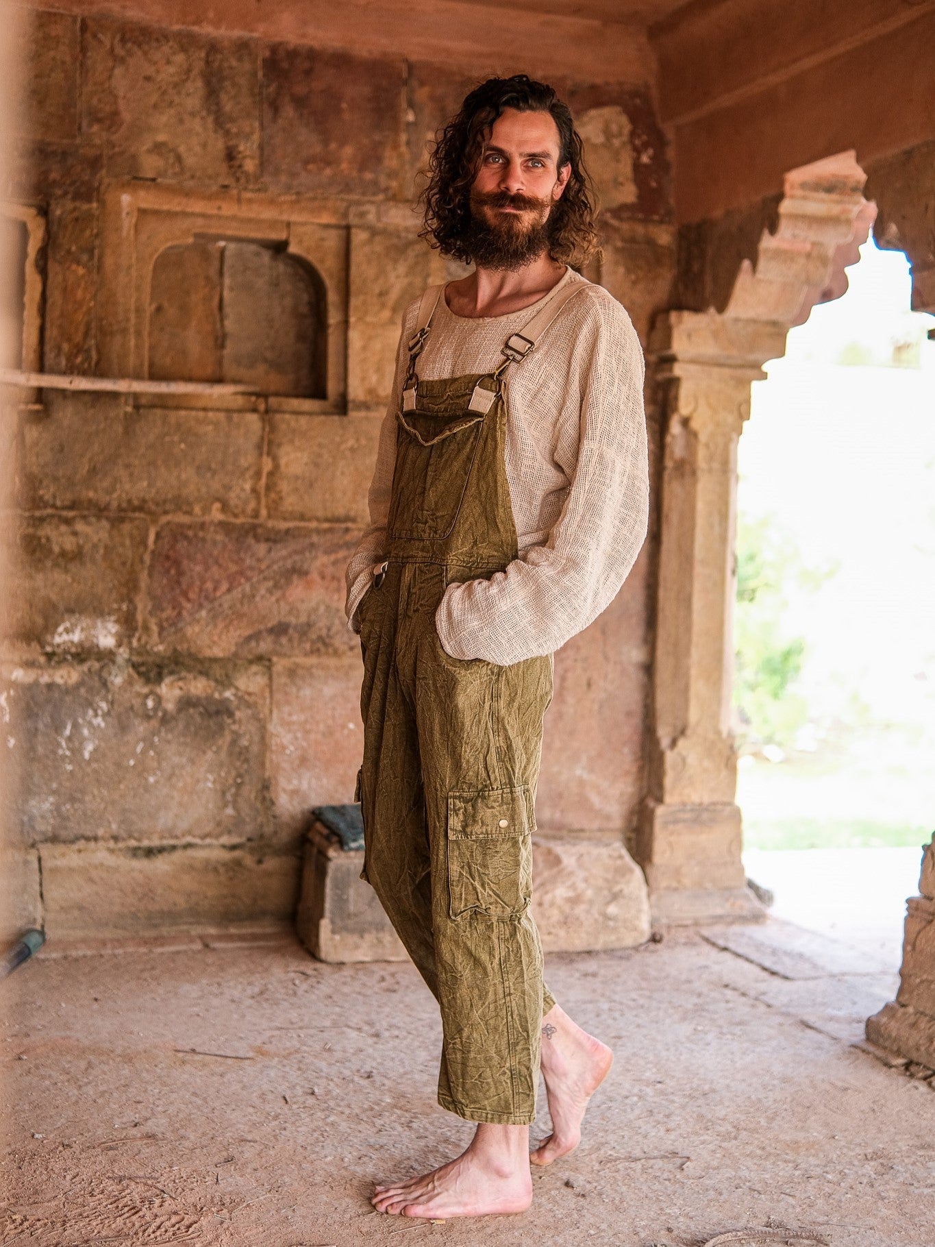 Man wearing a beige shirt and green overalls standing in a stone building with a bright light source.